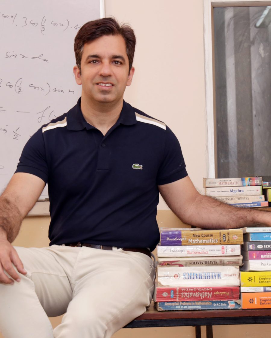 Sachin, founder of TruMath, sitting beside stacks of mathematics textbooks in front of a whiteboard covered in calculus working